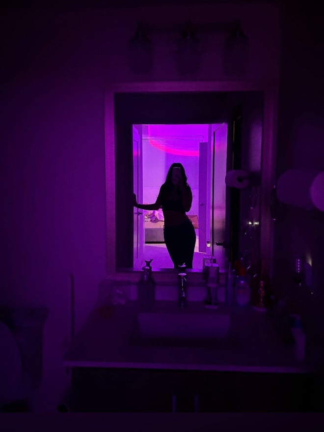 Silhouette of a person posing in front of a bathroom mirror with purple lighting and vanity items visible.