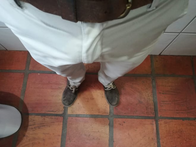 A person standing in white pants and brown belt on a tiled floor in a bathroom setting.