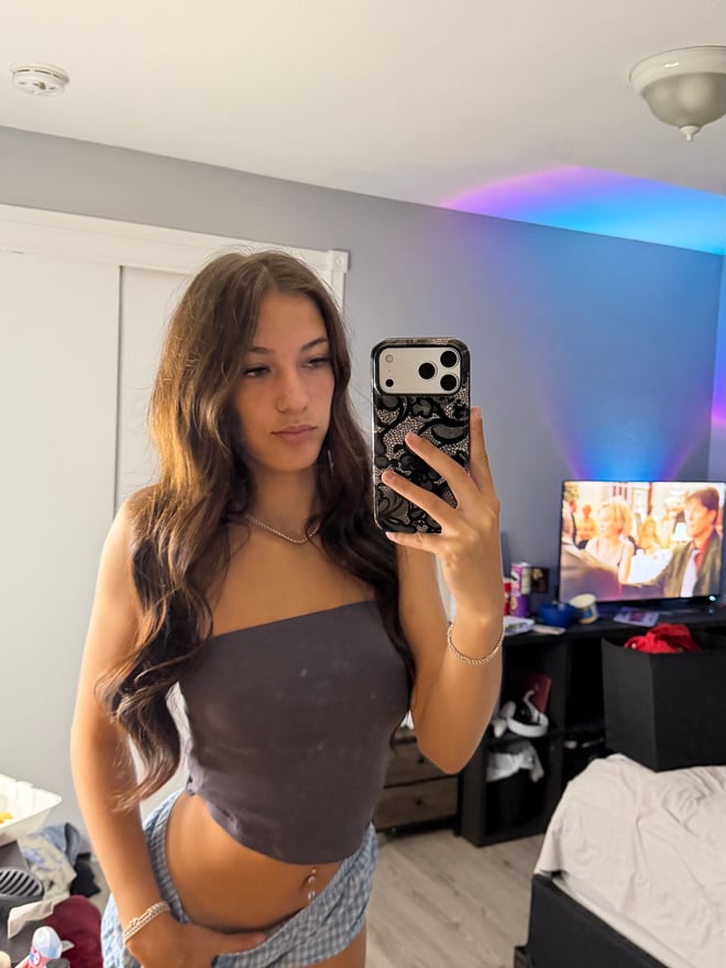 Young woman with long dark hair posing in a zebra-print bodysuit and fluffy boots, taking a mirror selfie in a bedroom with warm lighting.
