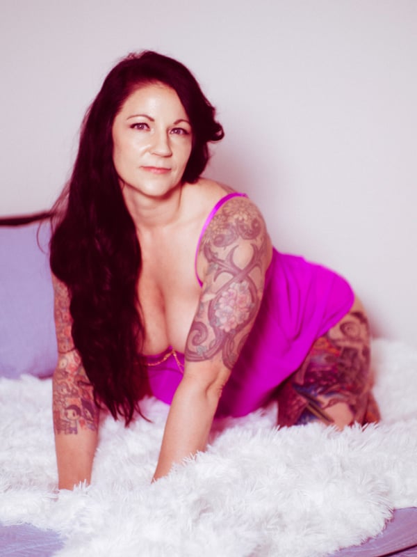 A woman with long brunette hair and tattoos, sitting on a bed in a pink top, smiling at the camera in a cozy bedroom setting.