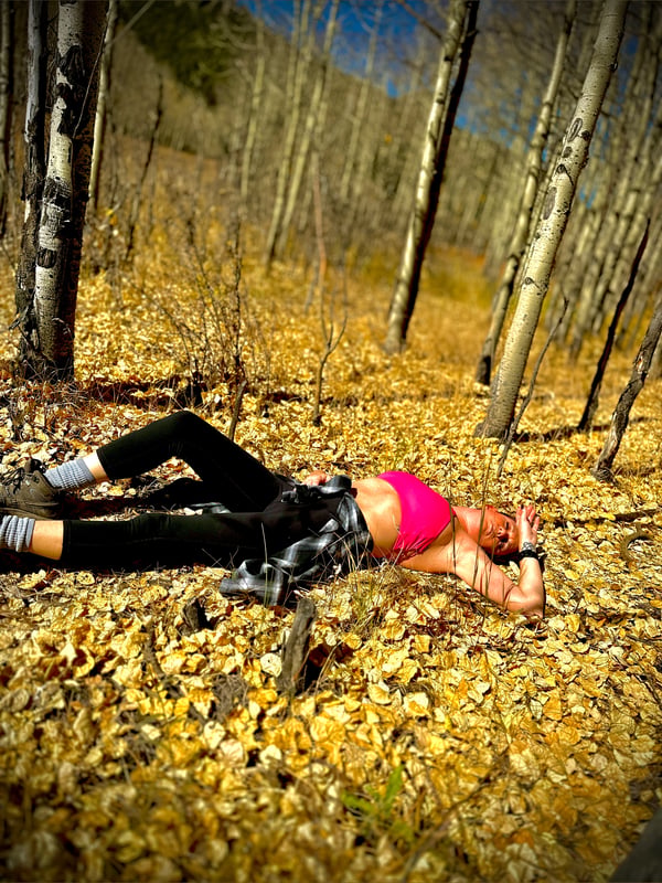 Woman lying on autumn forest floor surrounded by aspen trees and fallen leaves, wearing black leggings and a pink top.