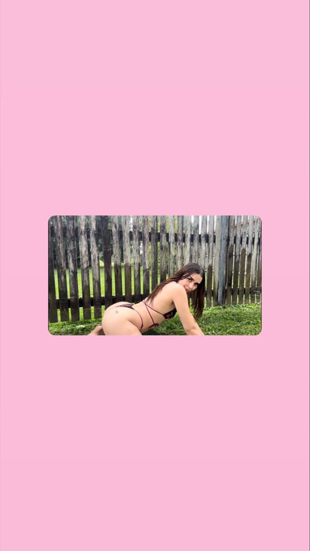 Young woman in a pink bikini posing playfully on grass near a wooden fence with a scenic outdoor background.