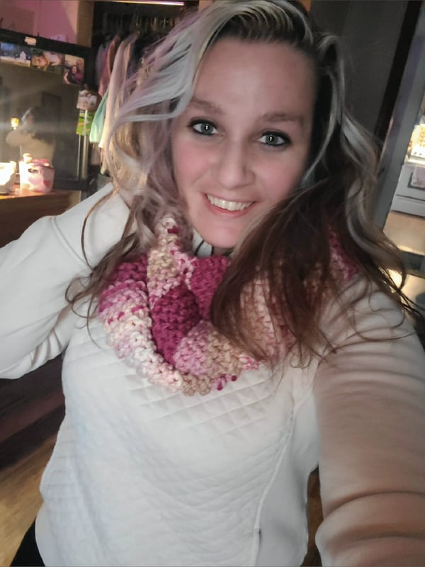 Young woman with highlighted hair smiling at the camera, wearing a white quilted top and colorful knitted scarf in a cozy indoor setting.