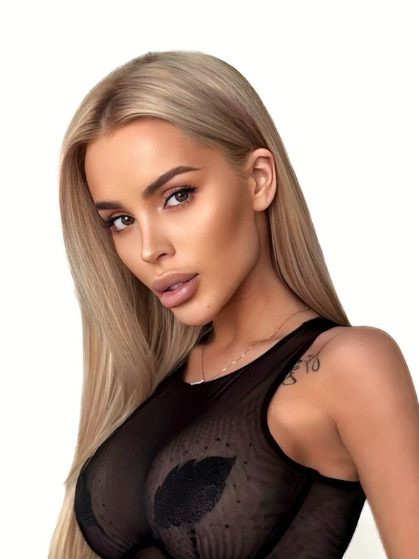 Close-up portrait of a young woman with long blonde hair, wearing a sheer black top, against a white background.