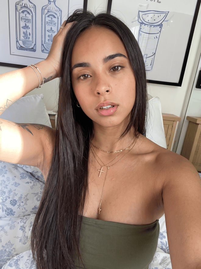 A young woman with long dark hair taking a casual portrait indoors, wearing layered necklaces and a strapless top.