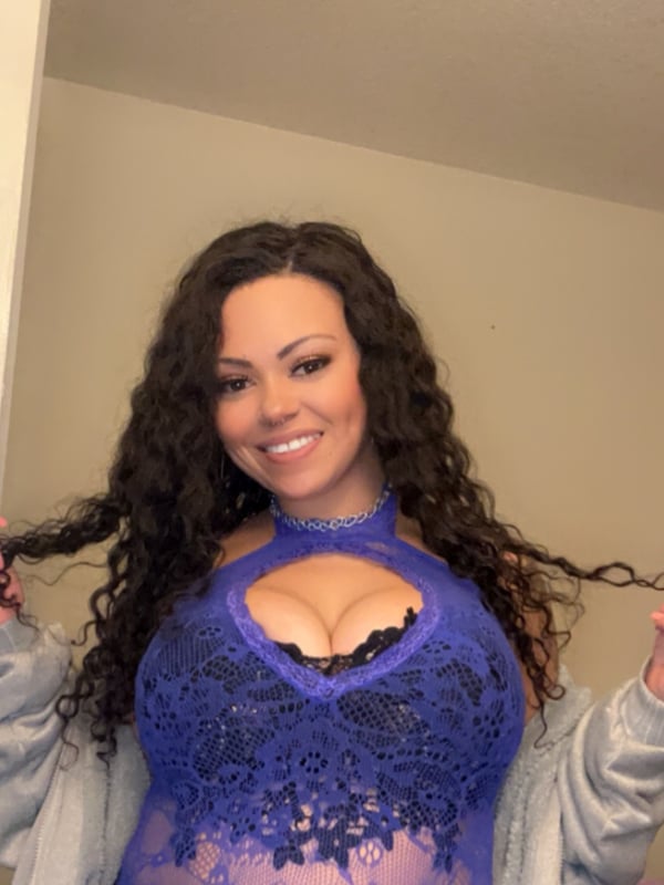Woman with long wavy dark hair smiling confidently while wearing a blue lace top and open cardigan indoors.
