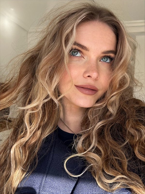 Close-up portrait of a young woman with wavy blonde hair, blue eyes, and subtle makeup, smiling softly against a neutral indoor background.