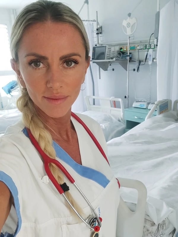 A blonde woman in a nurse uniform with a stethoscope, smiling in a hospital room with medical equipment.