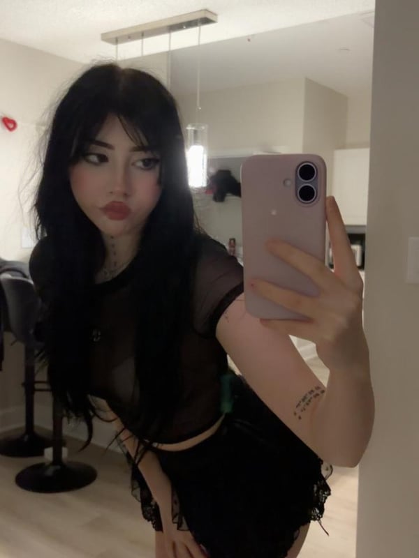 A person standing in a room wearing a black crop top and low-rise jeans, showing a navel piercing, with a wall outlet in the background.