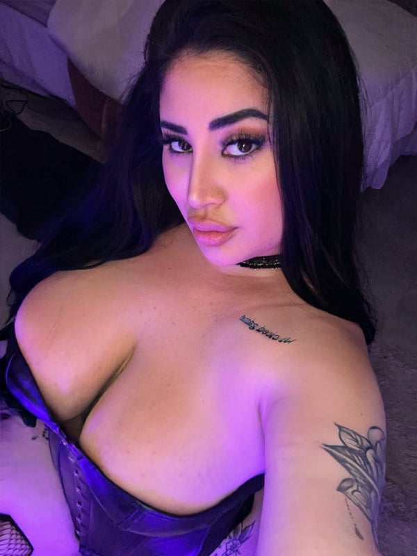 A woman with dark hair and makeup posing confidently in a low-cut purple top indoors, showcasing tattoos and a choker necklace.