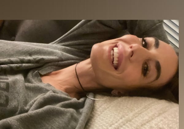 A smiling woman with long dark hair lying on a bed, wearing a gray top, with her head tilted back against pillows near window blinds.