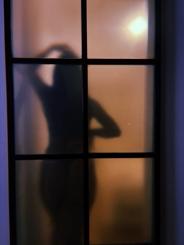 Silhouette of a woman with long hair standing behind a frosted glass shower door, illuminated by warm light.