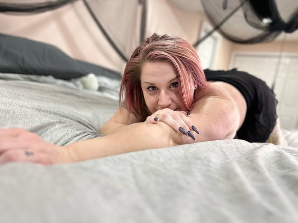 A woman with pinkish hair lying on a bed in a dimly lit room with draped fabric and dark bedding, posing playfully.