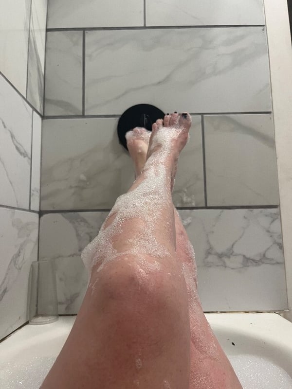 Close-up view of bare legs and feet positioned against a tiled shower wall with foam visible.
