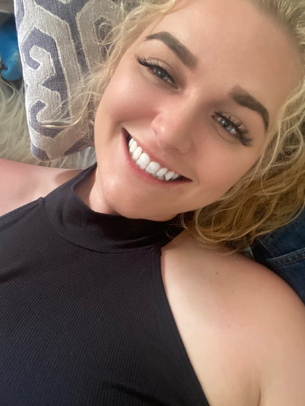 Young blonde woman with wavy hair smiling warmly while reclining on patterned pillows in a casual top.