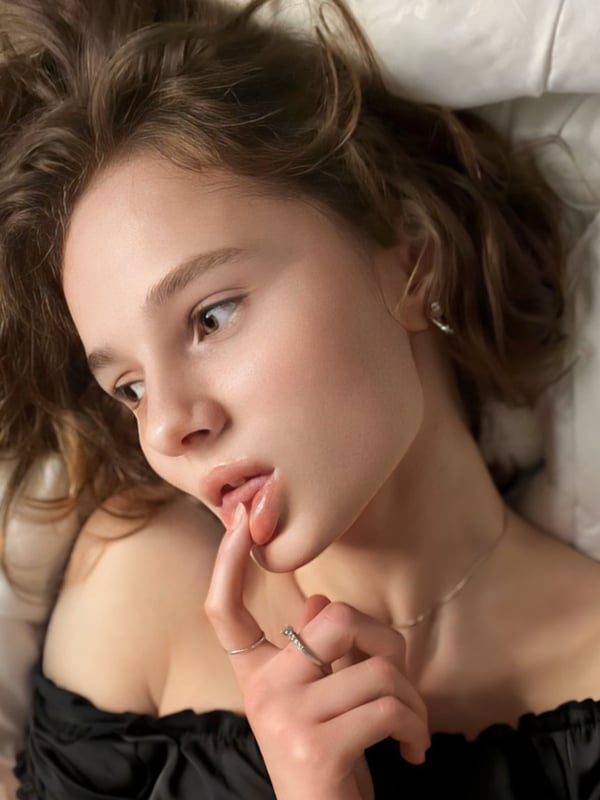Young woman with wavy brown hair lying on a bed, touching her lips thoughtfully while wearing a black off-shoulder top.