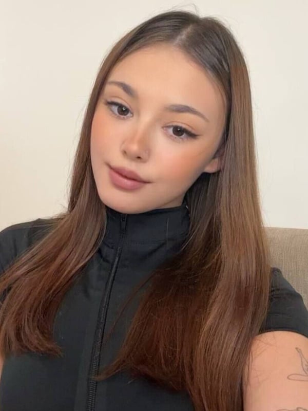Young woman with long brown hair taking a selfie in a white top, smiling softly against a plain background.