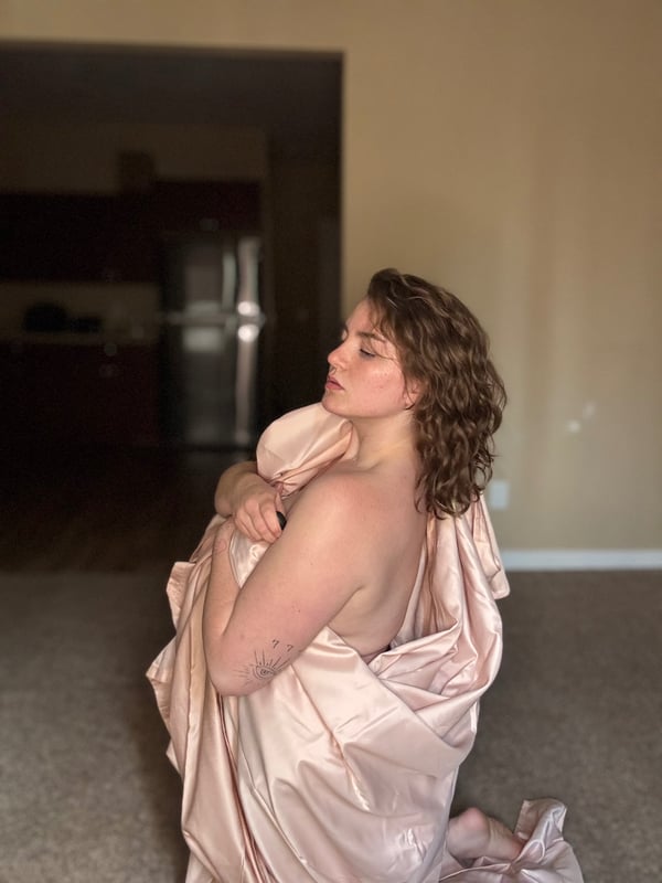 Young woman with curly brown hair kneeling on the floor in a home setting, draped in a pink satin sheet.
