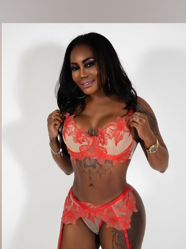 A woman posing confidently in red lace lingerie against a gray background, showcasing tattoos on her arms and legs.