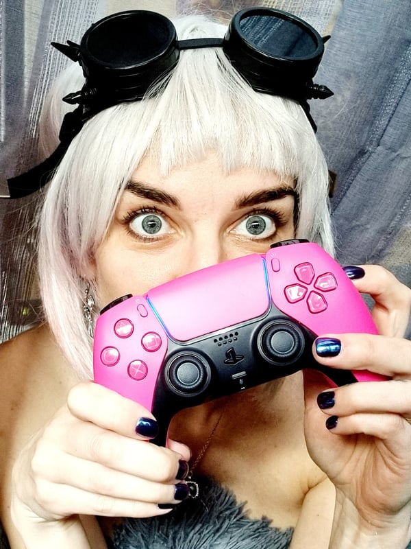 A woman with platinum blonde hair and steampunk goggles holding a pink gaming controller, smiling playfully in front of a curtain.