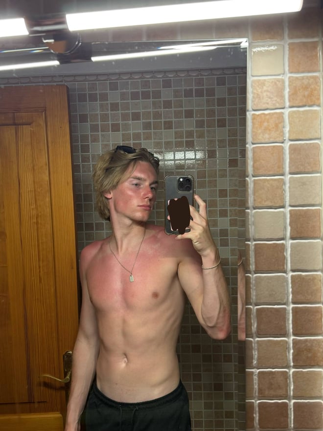 Young man with curly blonde hair taking a mirror selfie in a locker room, wearing a white t-shirt and grey sweatpants.