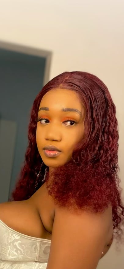 A young woman with long wavy red hair posing confidently indoors, wearing a white off-shoulder top.