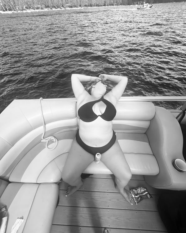 Woman lounging on a boat deck in black swimwear with arms raised, overlooking a scenic lake and distant shoreline in black and white.