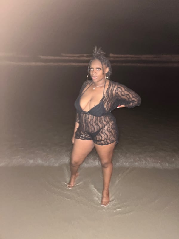 A woman standing in shallow beach water at night, wearing a sheer black outfit.