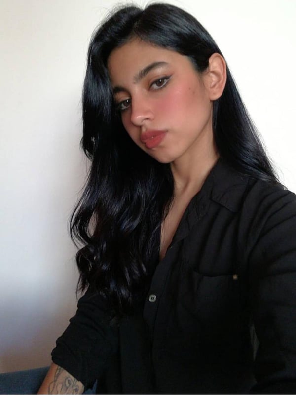 Close-up portrait of a woman with long dark hair, wearing a black shirt, with subtle makeup and tattoos on her arm.