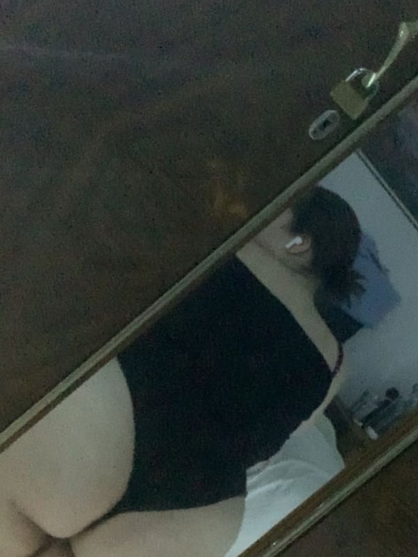 A person with dark hair standing in front of a mirror in a room, wearing a black swimsuit and showing their backside.
