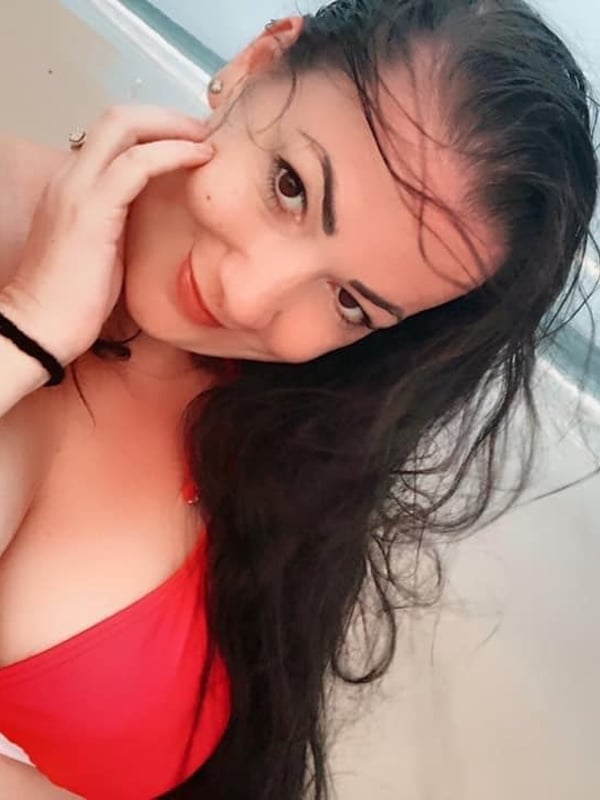 Woman with long dark hair smiling at the camera, wearing a red swimsuit top while sitting by the beach.