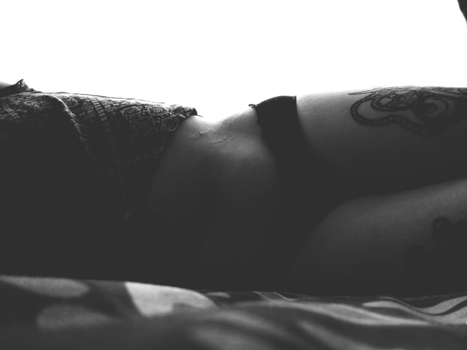 A person lying on their side in dim lighting, wearing torn clothing and displaying arm tattoos.