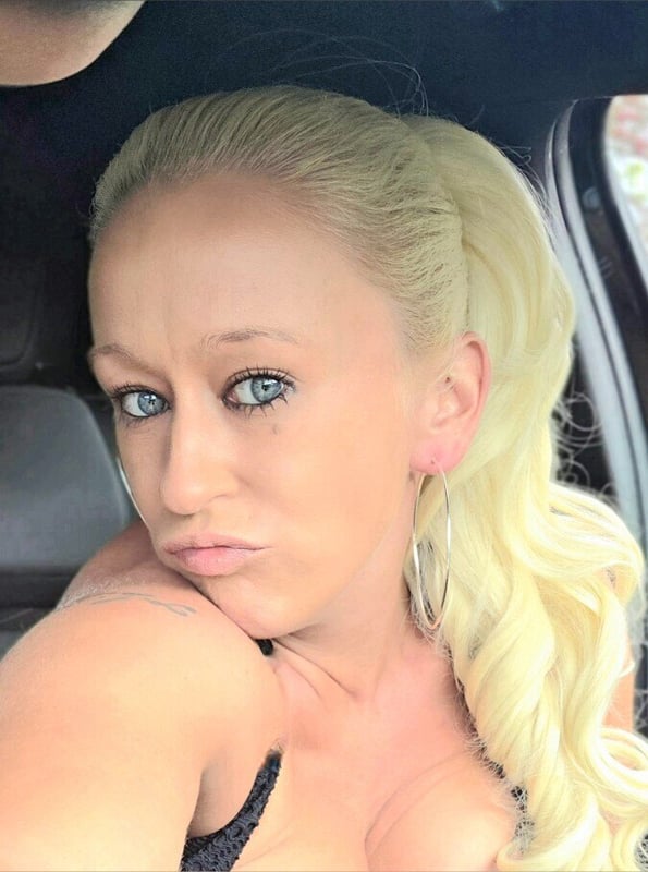 A blonde woman with a ponytail posing with a pout in a car, wearing a black top.