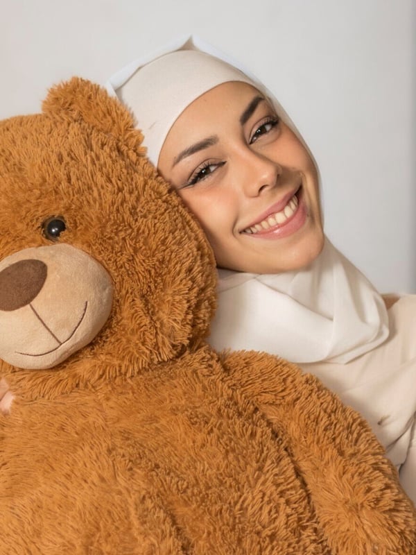 Smiling young woman in a white hijab hugging a large teddy bear against a plain background.
