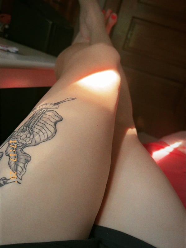 Close-up of a person's bare legs and feet with a detailed tattoo on the thigh, posed indoors near a door.