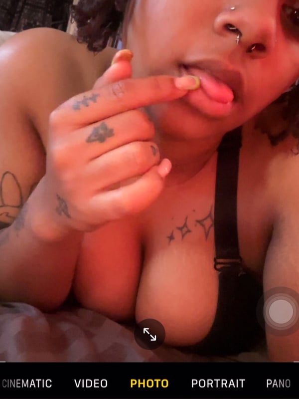 Close-up portrait of a person with curly hair, piercings, and tattoos, posing playfully with tongue out while wearing a top.