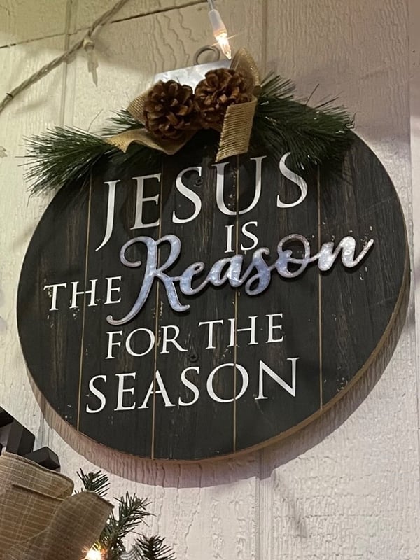 A wooden holiday sign reading 'Jesus is the Reason for the Season' decorated with pine branches, pinecones, and gold ribbons, hanging against a white wall with festive lighting.