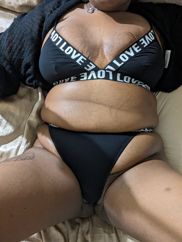 A woman lounging on a bed in black lingerie, posing relaxed with arms extended.