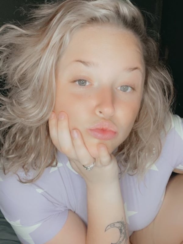 A woman with wavy blonde hair smiling softly while resting her chin on her hand, wearing a light-colored top in a cozy indoor setting.