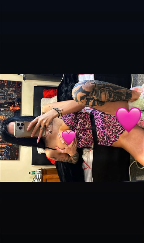 A woman with long dark hair and tattoos poses playfully on a bed, holding heart emojis over her chest while wearing a colorful leopard print outfit in a cozy bedroom setting.