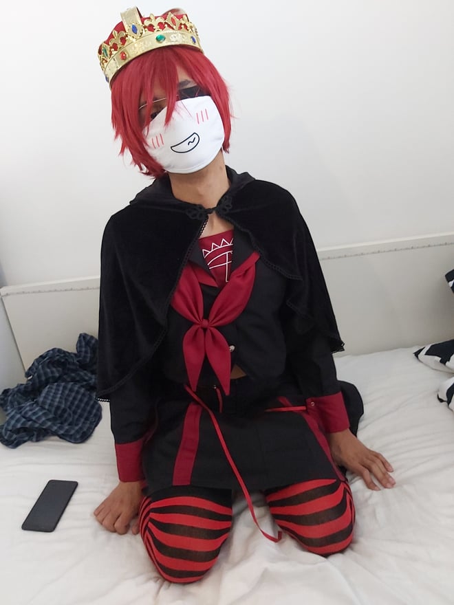 A person in a black and red hooded costume with a crown and mask, sitting on a bed with striped stockings.