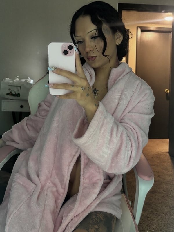 A woman with long dark hair taking a selfie in a pink robe inside a bedroom, holding her phone with manicured nails.
