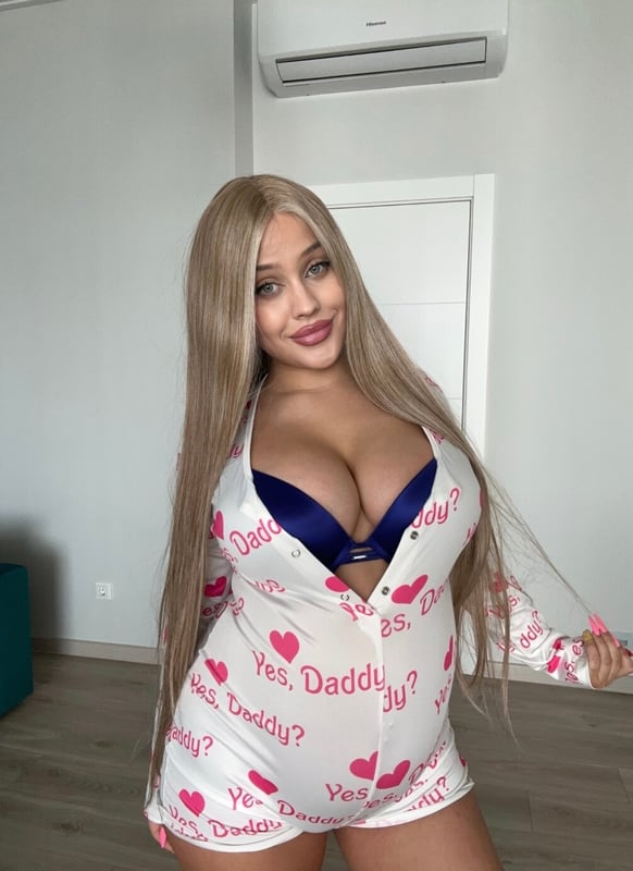A woman with long blonde hair posing indoors in a white patterned outfit over a blue top.