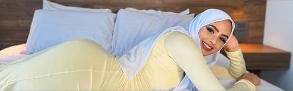 A woman wearing a white hijab and yellow top poses on a bed in a cozy bedroom with wooden accents and pillows.