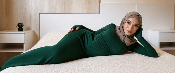 Woman in a green hijab and form-fitting green dress lounging on a bed in a modern bedroom with wooden paneling and bedside tables.