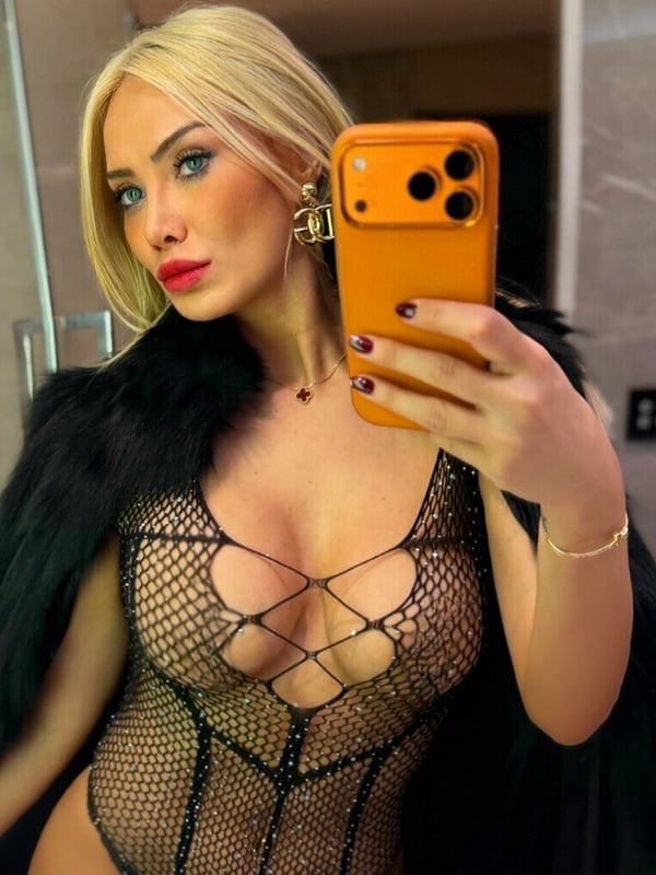 A woman with long blonde hair taking a mirror selfie in a bathroom, holding an orange smartphone, dressed in a sparkling black outfit with fur accents.