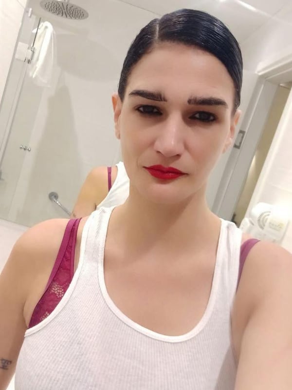 A woman with dark hair and bold makeup stands in a bathroom mirror selfie, wearing a white tank top with red straps, with tiled walls and a shower in the background.