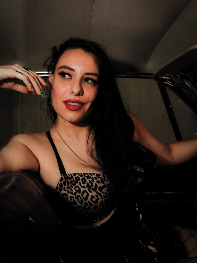 A woman with long dark hair posing playfully in a car interior, wearing a leopard print top and smiling at the camera.