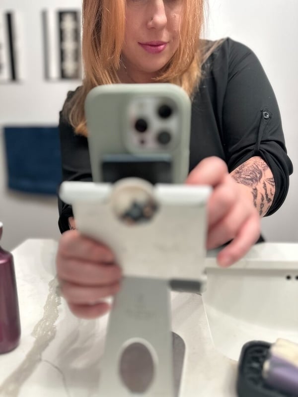A woman with long blonde hair holding her green iPhone in a bathroom setting, wearing a black top, with tattoos visible on her arm.