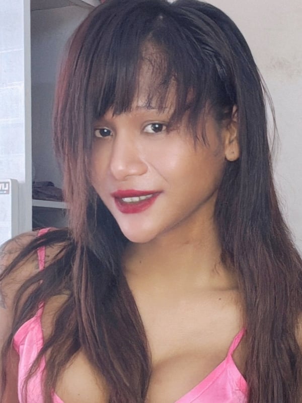 Young woman with long dark hair posing indoors wearing a pink top, smiling at the camera with decorative elements in the background.
